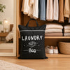 A black laundry bag with a handle is displayed on a wooden shelf among folded textiles.