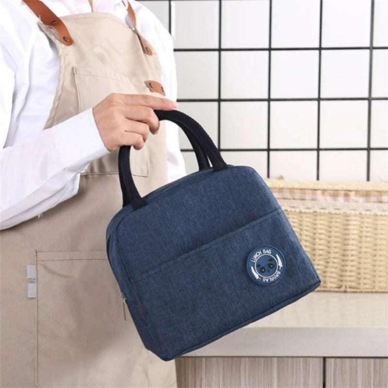 A hand holds a dark blue insulated lunch bag with black handles against a light beige apron.