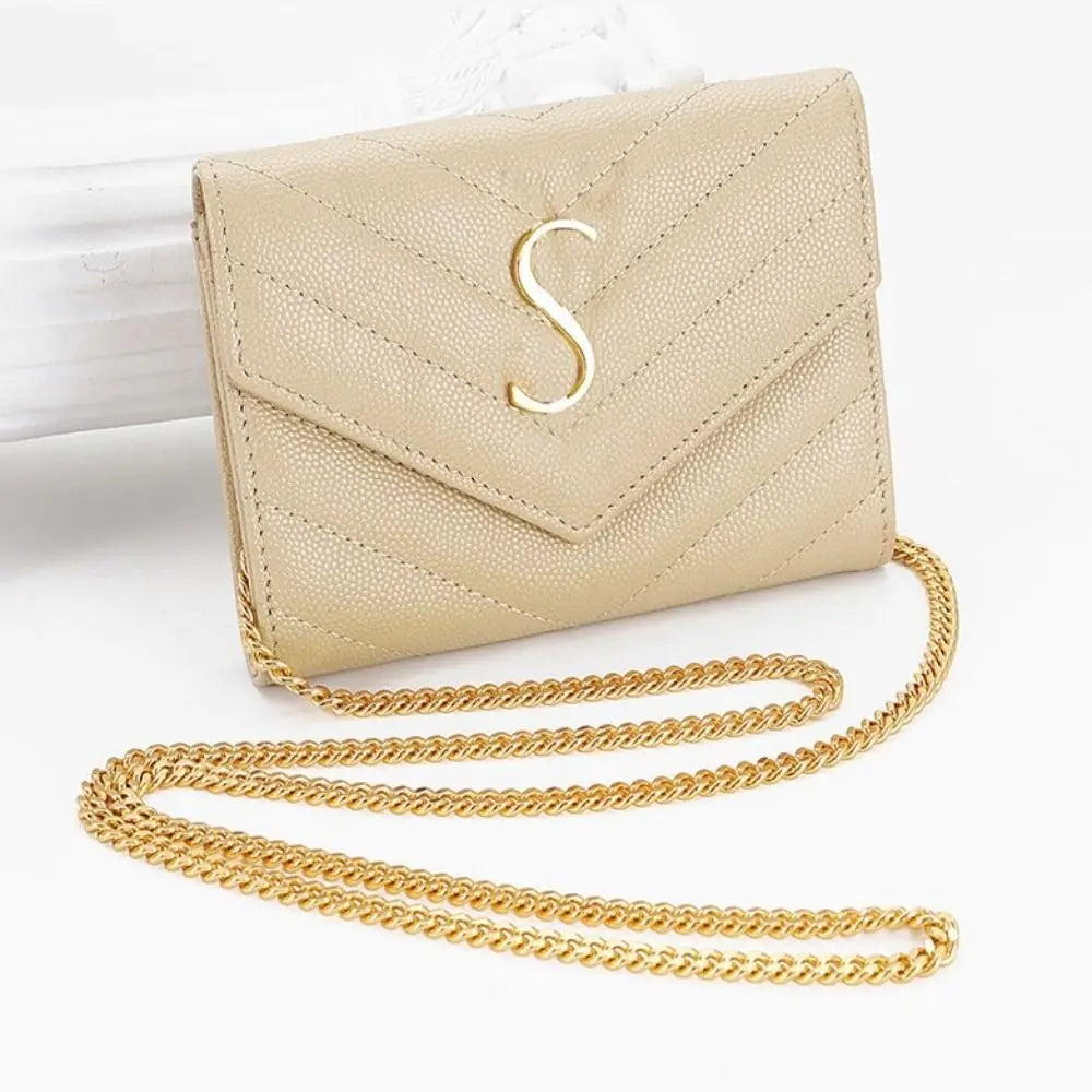A beige quilted leather clutch with a gold chain strap and a silver initial S detail.