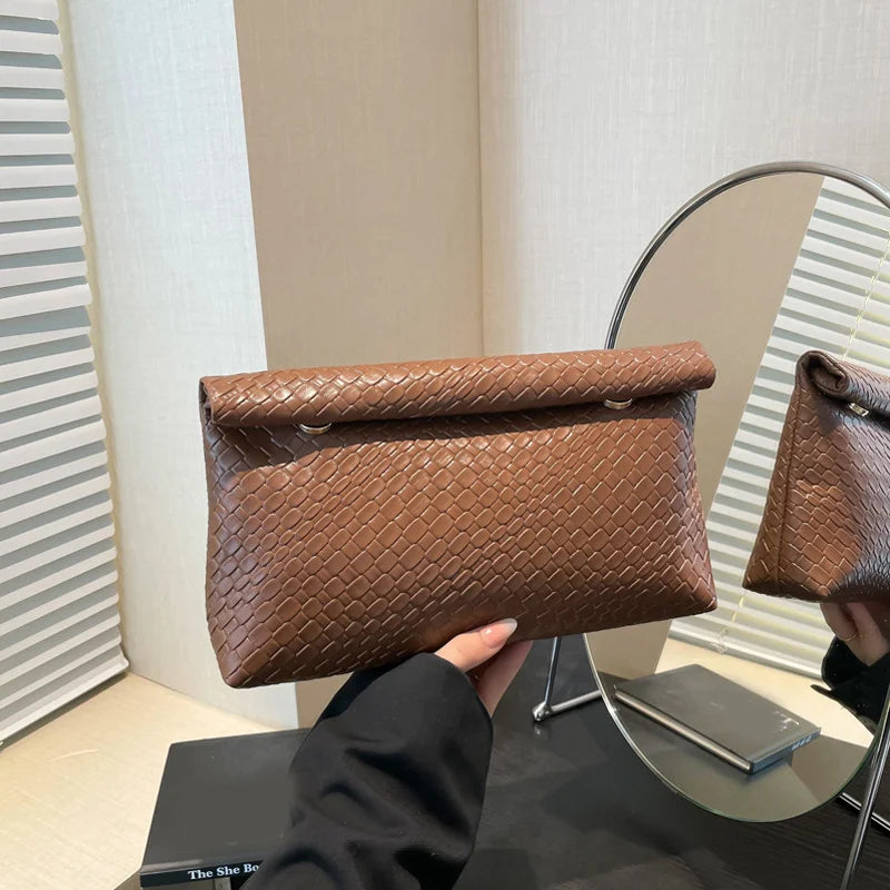 Un bolso de mano tejido marrón se muestra sobre una superficie de cristal junto a un espejo de marco plateado.