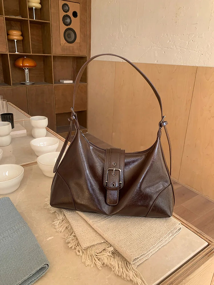 A brown retro PU leather underarm bag with a long strap rests on a light surface next to a beige fabric.