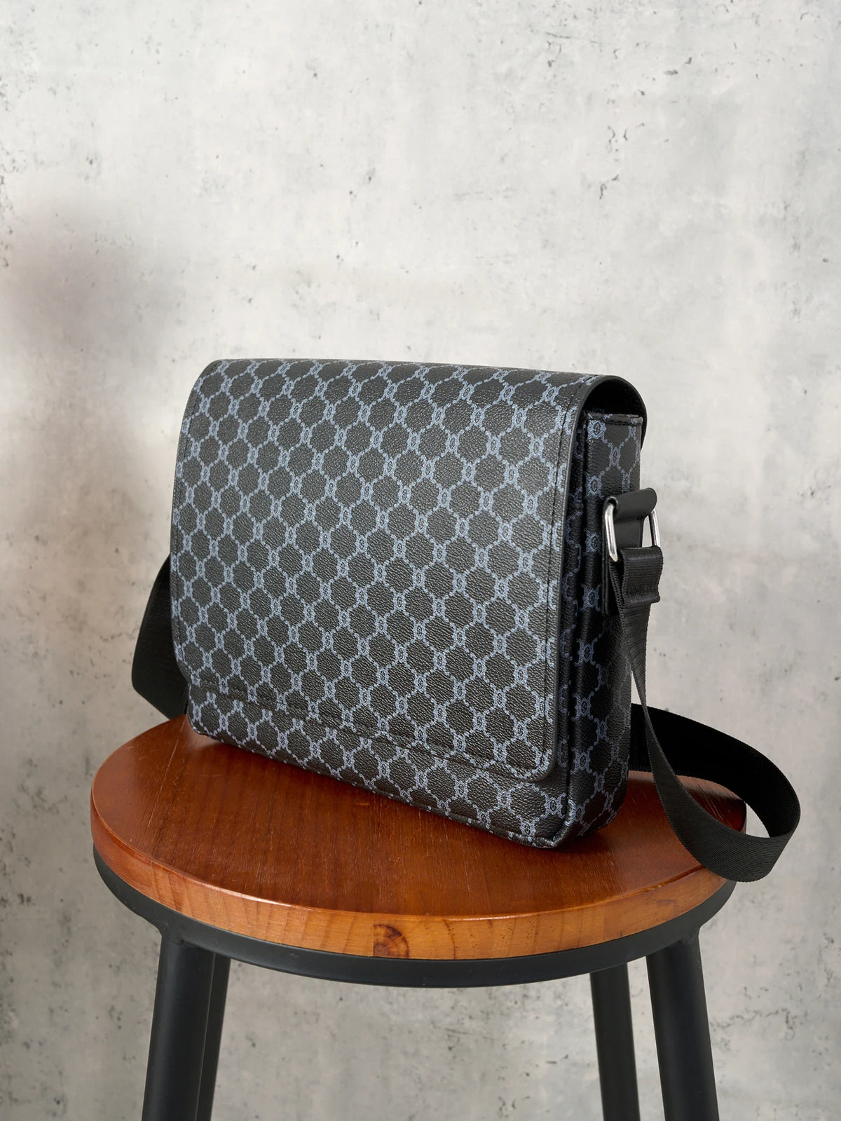 A dark gray patterned flap crossbody bag resting on a wooden stool.