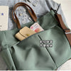 A light sage green nylon tote bag with brown leather straps and a flap pocket, shown against a denim background.