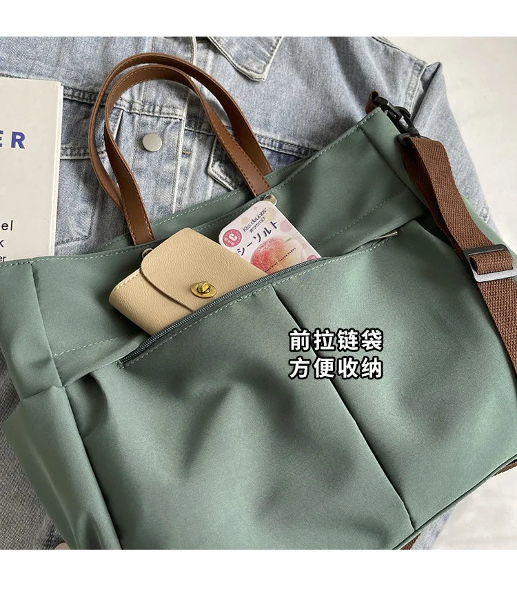 A light sage green nylon tote bag with brown leather straps and a flap pocket, shown against a denim background.