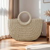 A large, light beige handwoven rattan beach bag resting on a wooden floor next to a window.