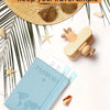 A light blue slim passport holder is shown next to a straw hat, sunglasses, and starfish on a white background.