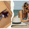 A woman wearing sunglasses and a head wrap sits on the beach with a foldable mesh tote bag nearby.