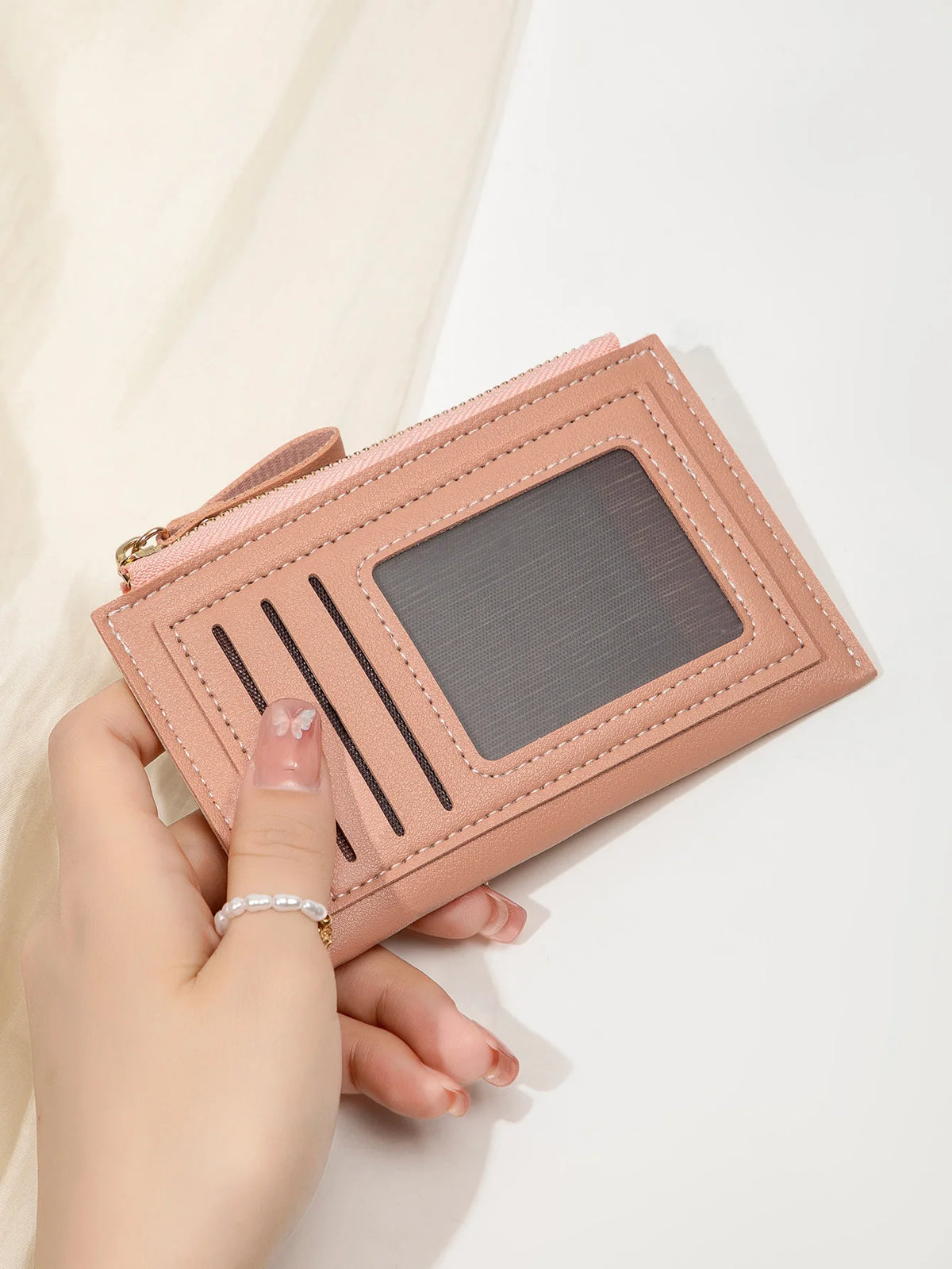 Une main tient un portefeuille en cuir PU rose minimaliste avec une fenêtre transparente et des rayures noires.