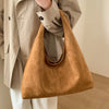 A tan suede shoulder tote bag carried by a person wearing a beige jacket and white pants.