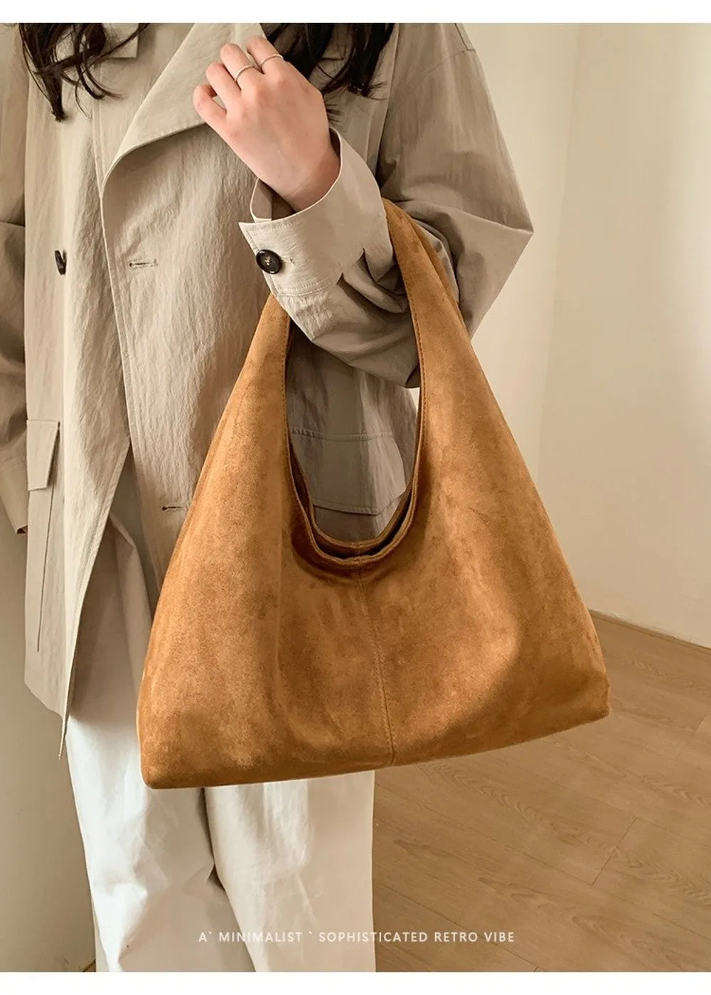 A tan suede shoulder tote bag carried by a person wearing a beige jacket and white pants.