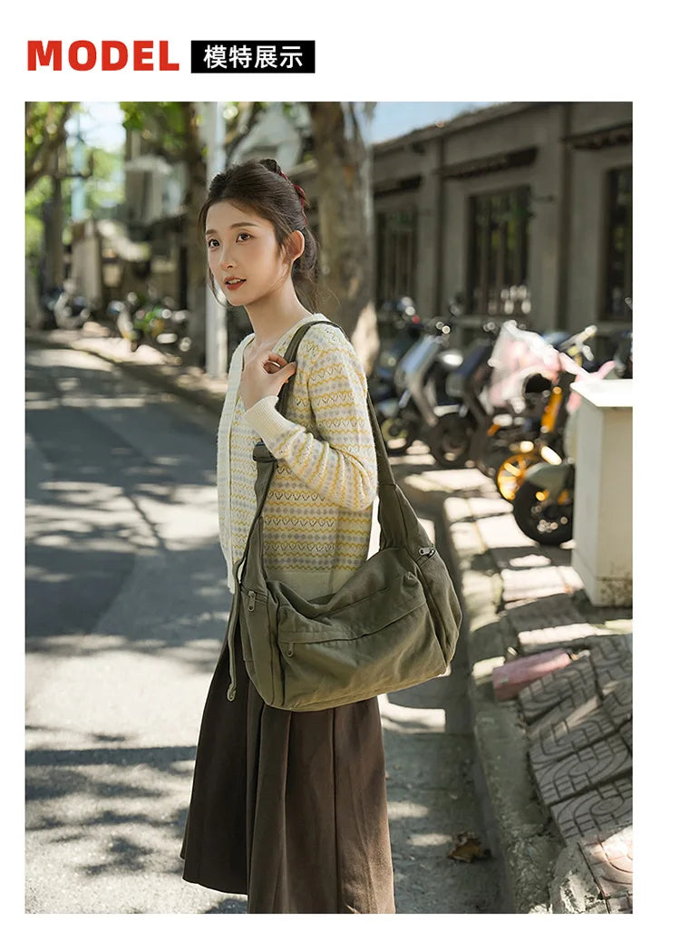 A young woman wearing a light yellow sweater and dark skirt stands on a sidewalk with a large olive green cotton canvas shoul