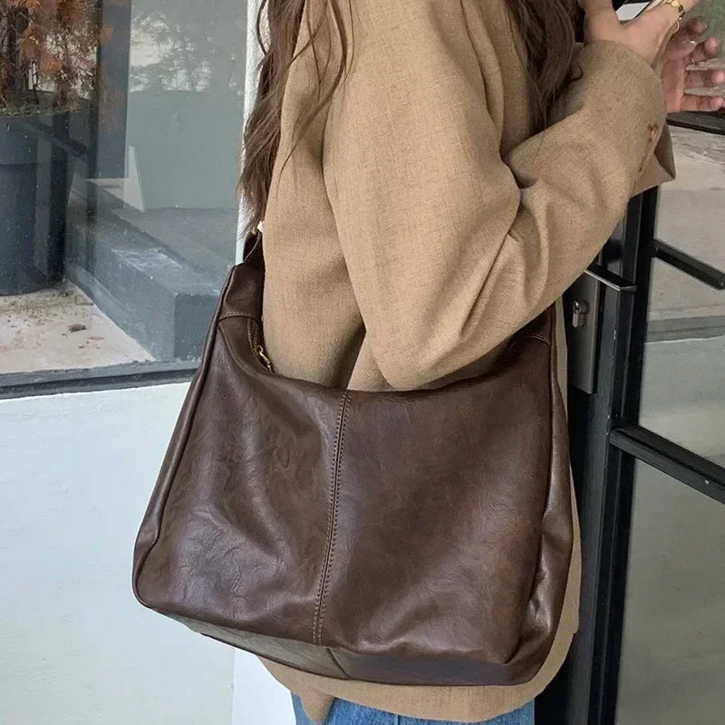 A person is holding a brown vintage PU leather tote bag with a crossbody strap against a glass window.
