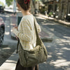 A woman wearing a cream knit sweater and dark skirt carries a large olive green cotton canvas shoulder crossbody bag on a cit
