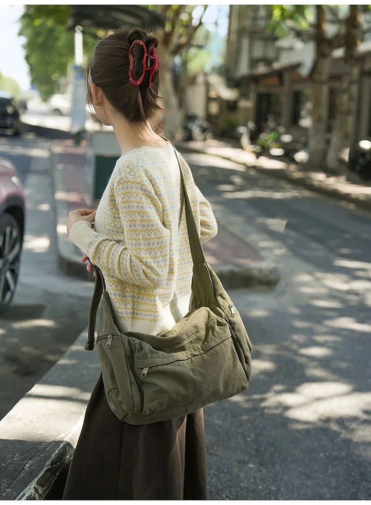 A woman wearing a cream knit sweater and dark skirt carries a large olive green cotton canvas shoulder crossbody bag on a cit