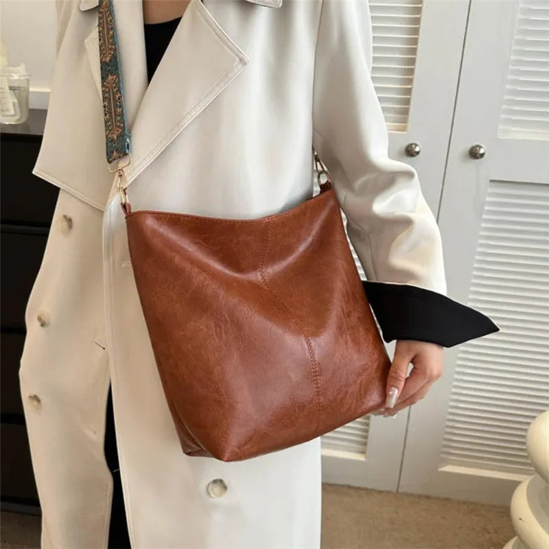A woman holds a brown leather shoulder bag with an embroidered detail against a white coat.
