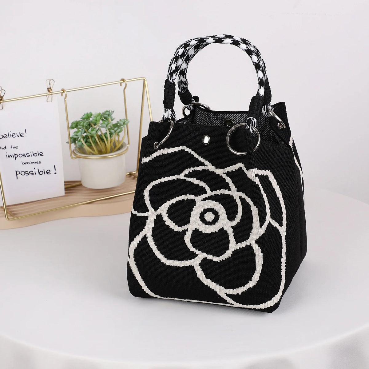 A black knitted bucket bag with a white geometric floral pattern and black and white striped handles sits next to a small pot