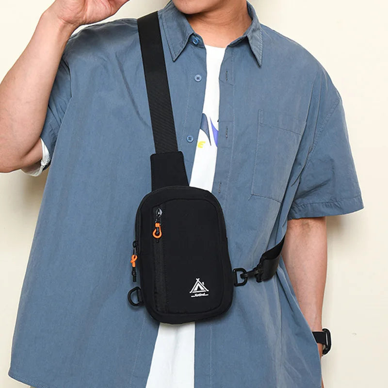 A person wearing a light blue shirt is holding a black nylon sling crossbody bag with an orange strap.