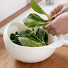A hand is holding a leafy green plant in a white plastic drain and storage basket on a wooden surface.