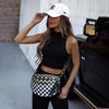 A woman wearing a black top and pants holds a checkered nylon crossbody bag while standing next to a dark car.