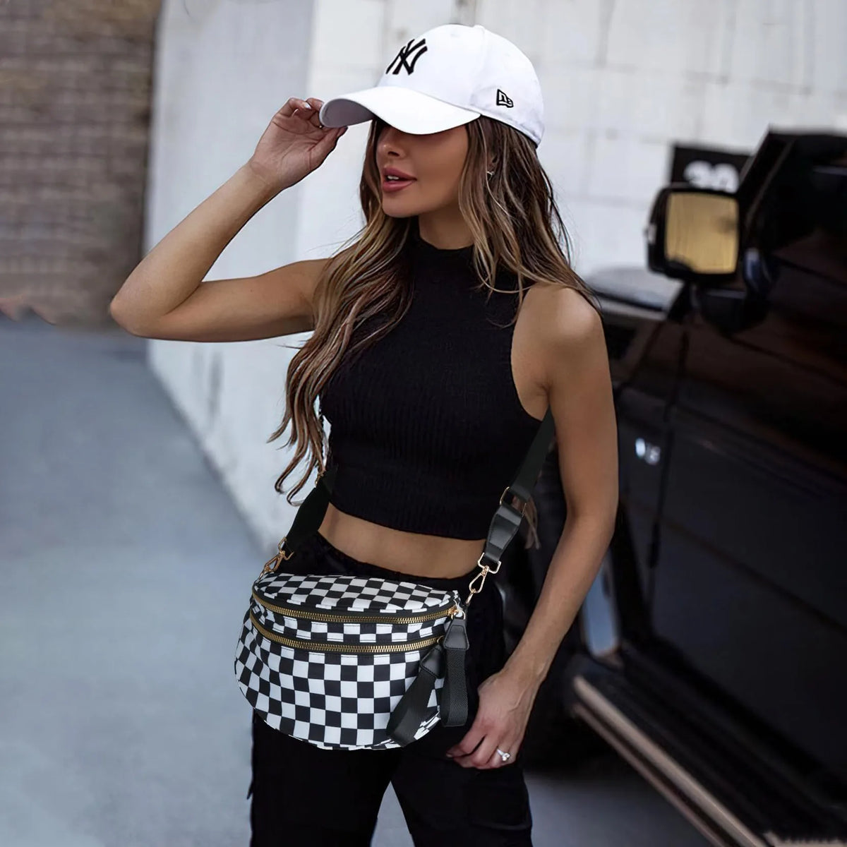 A woman wearing a black top and pants holds a checkered nylon crossbody bag while standing next to a dark car.