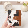 A hand holds a black and white cow print compact wallet with a brown leather strap on a white surface.