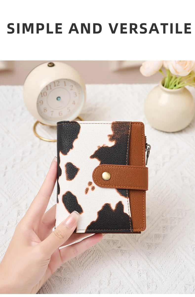 A hand holds a black and white cow print compact wallet with a brown leather strap on a white surface.