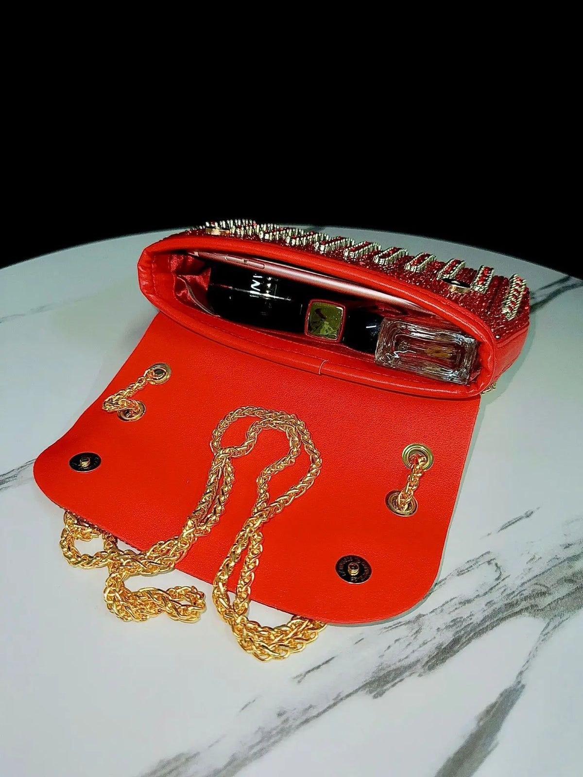 A red evening clutch bag adorned with rhinestones and featuring a diamond chain resting on a white marble surface.