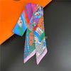 A colorful silky twill ribbon scarf featuring intricate patterns in pink, blue, and green, displayed against an orange box.