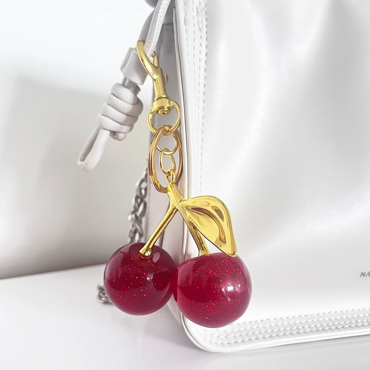 A gold-colored crystal cherry fruit keychain attached to a white leather handbag.