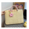 A light yellow quilted laptop sleeve with a pink handle and decorative charm is displayed on a black and white checkered surf
