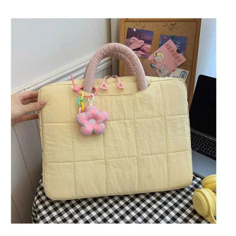 A light yellow quilted laptop sleeve with a pink handle and decorative charm is displayed on a black and white checkered surf