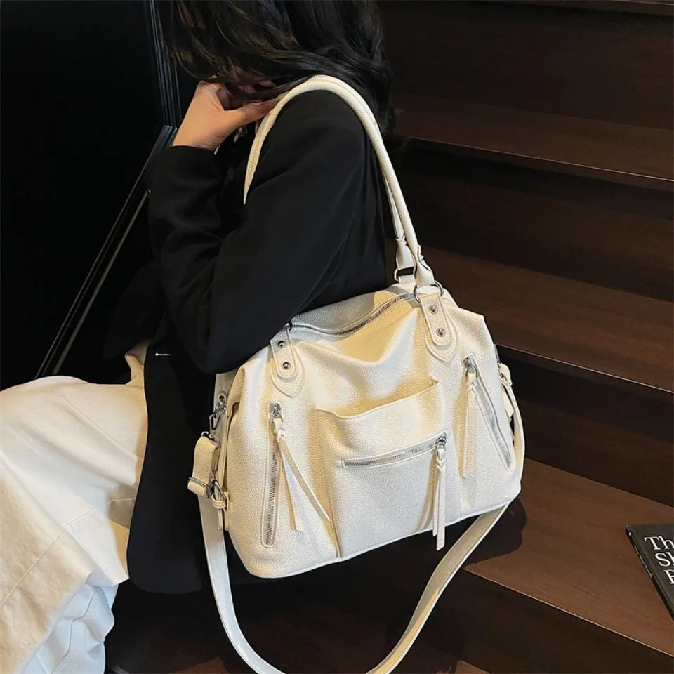 Una mujer sostiene un bolso cruz de cuero suave color crema contra un fondo oscuro.