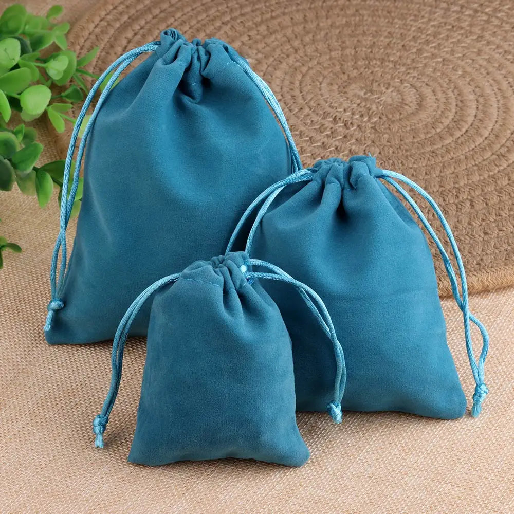 Un juego de tres bolsas de regalo de terciopelo verde azulado con cordones sobre una superficie tejida beige.