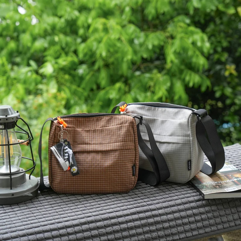 A tan and gray water resistant crossbody shoulder bag resting on a dark gray textured surface with green foliage in the backg