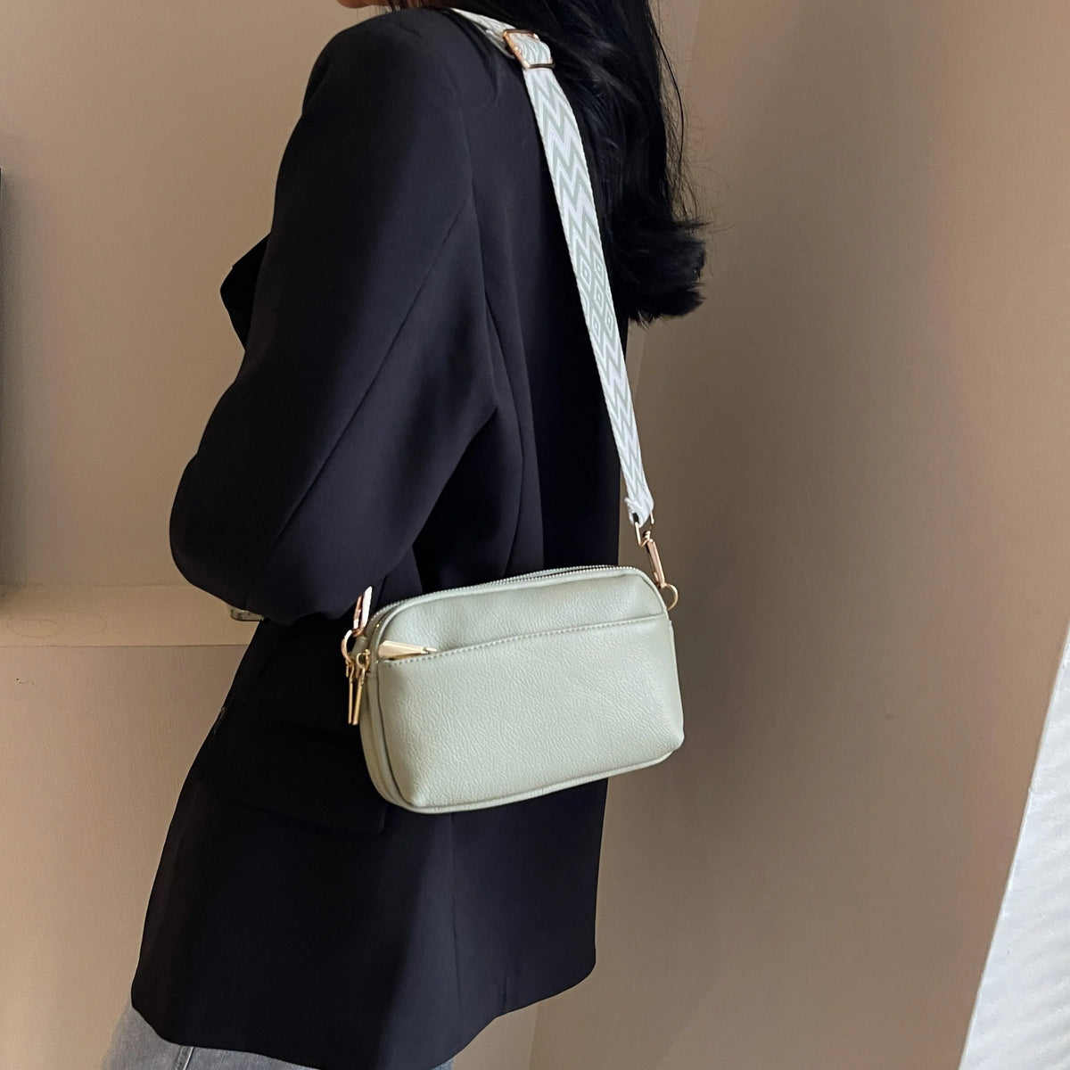 A woman is holding a light green vegan PU crossbody bag with a white striped strap against a neutral background.