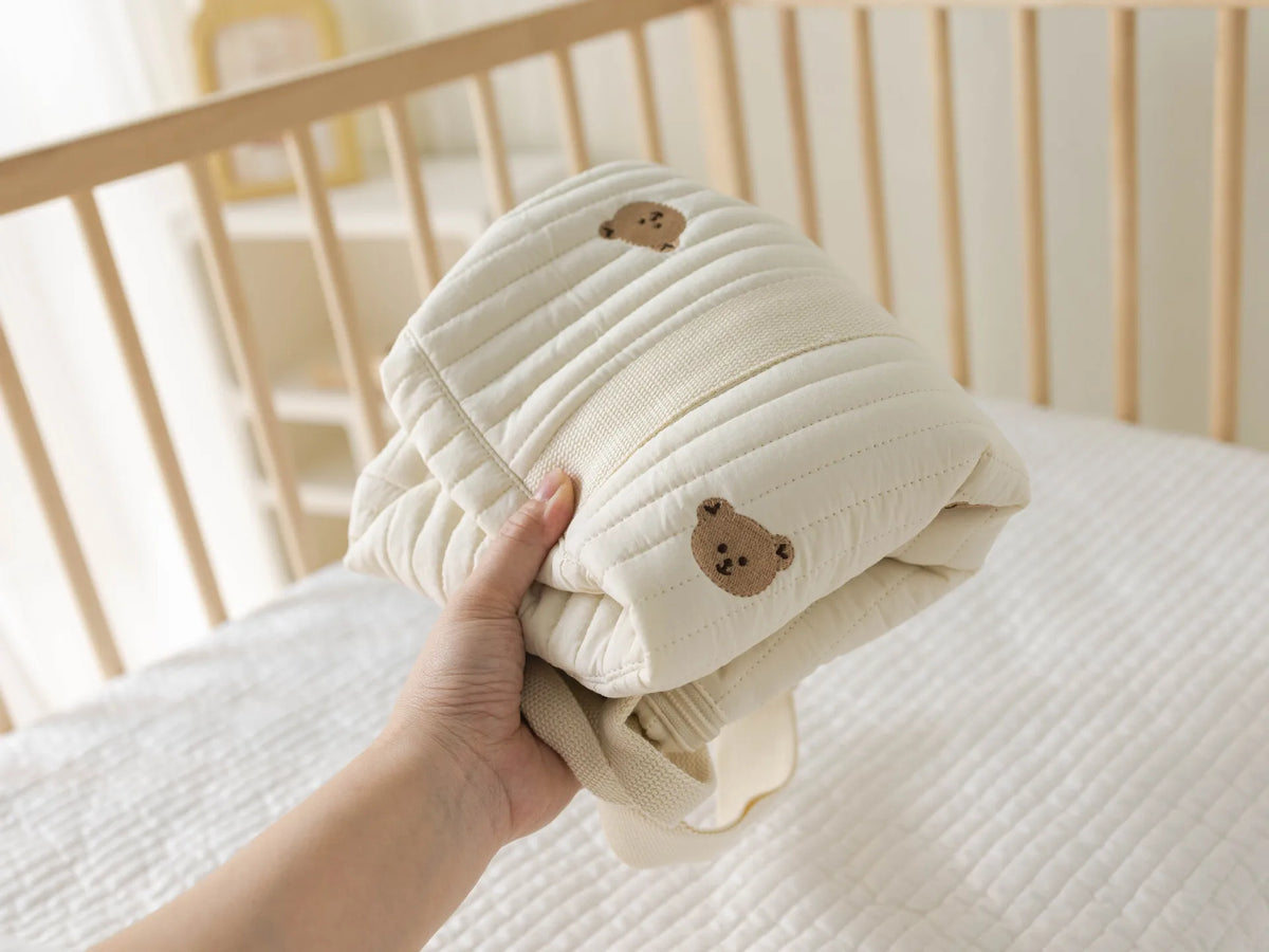 A hand is holding a cream-colored maternity and baby diaper bag tote with brown bear designs.