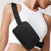 A close-up of a black cross-body nylon waist bag worn across the chest of a woman wearing a white ribbed tank top.