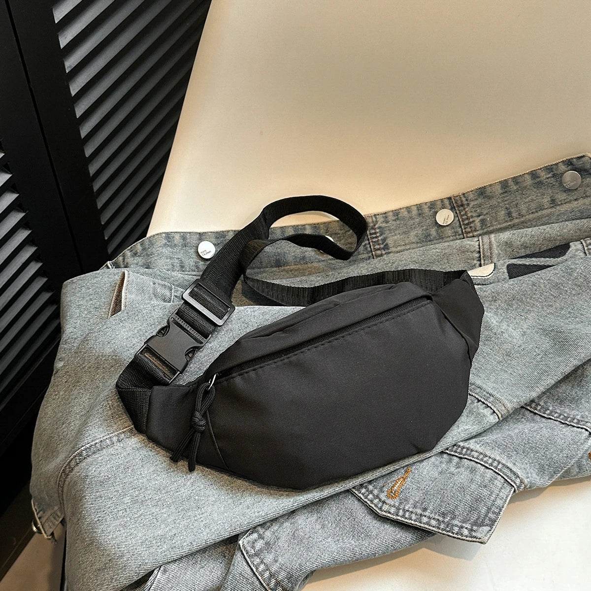 Un sac bandoulière noir en nylon est exposé sur une veste en denim gris.
