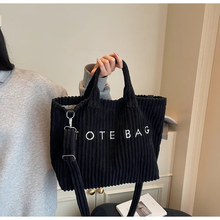 Un sac à épaule en velours rayé noir est tenu par une personne dans un intérieur bien éclairé.