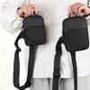 A black canvas crossbody sling bag is being held by a person against a white shirt.
