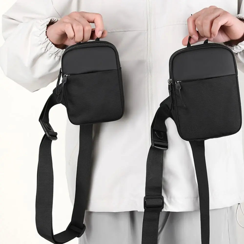 A black canvas crossbody sling bag is being held by a person against a white shirt.