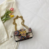A small patterned crossbody bag with gold chain strap rests on a white sheet next to pink and yellow roses.