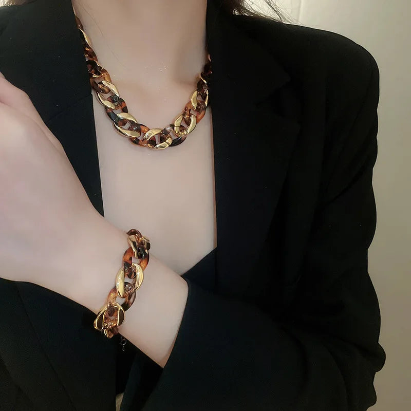 A close-up of a woman wearing a gold leopard print interlocking dangle necklace and bracelet under a black blazer.