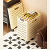 A cream-colored foldable laundry basket with a perforated design is shown on a wooden floor next to a lamp and a brown leathe