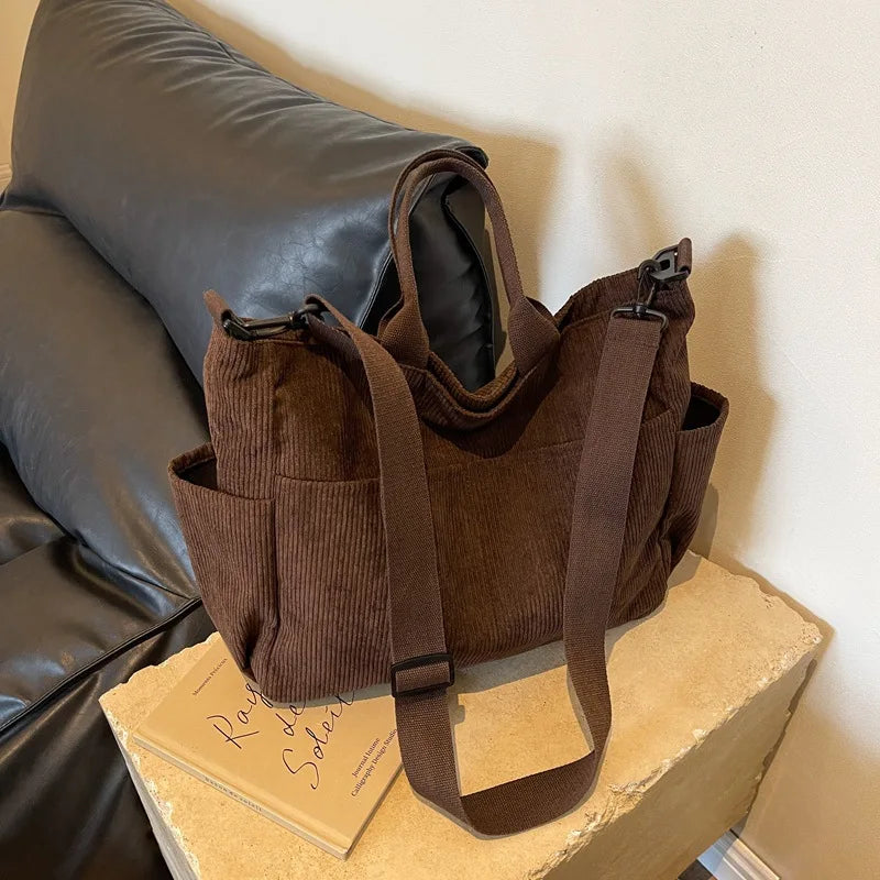 A brown corduroy multi-pocket tote bag resting on a light wooden surface next to a dark leather cushion.