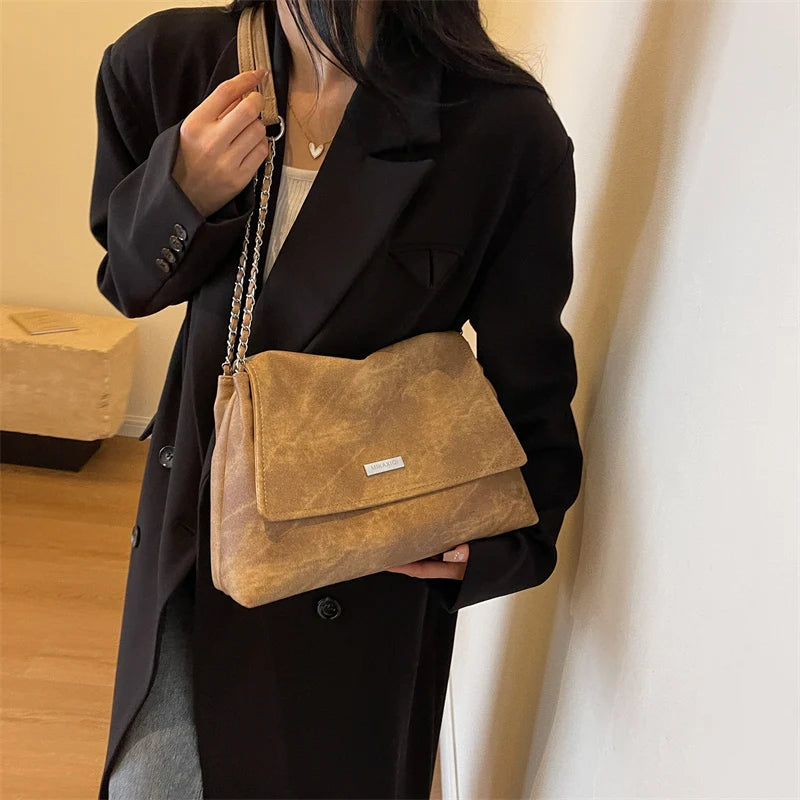 Una mujer sostiene un bolso de cuero color bronce con correa de cadena dorada sobre un fondo blanco.