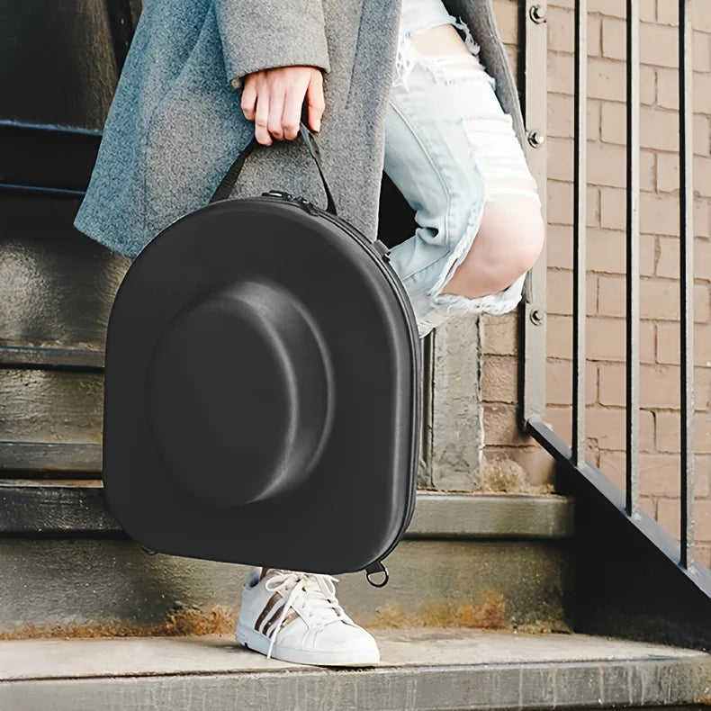 A black durable EVA hard shell hat case is resting on a concrete ledge next to a person wearing light-wash jeans and a gray c