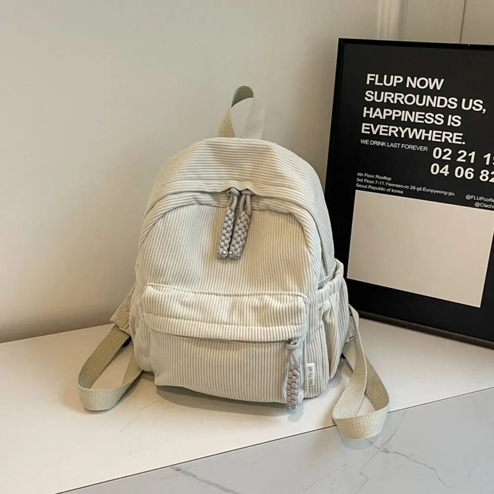 A light beige lightweight corduroy daily backpack is displayed on a white surface next to a framed sign.