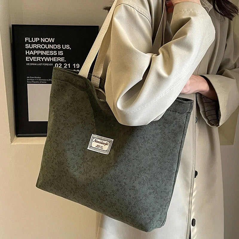 A light beige jacket sleeve holds a dark green bohemian corduroy tote bag against a white wall.