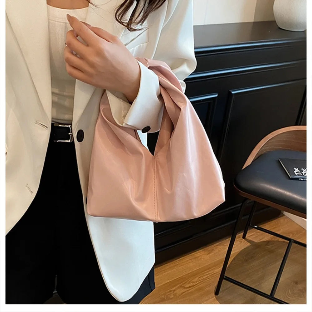 A woman holds a light pink leather minimalist crossbody tote bag against a white blazer and black pants.